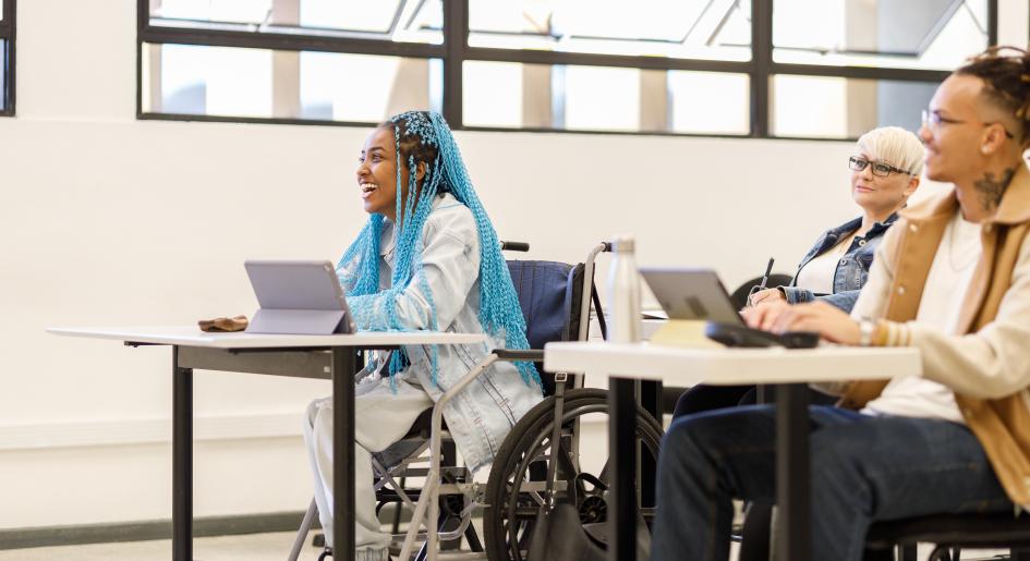 A group of students sit in a classroom. In the center is a wheelchair user